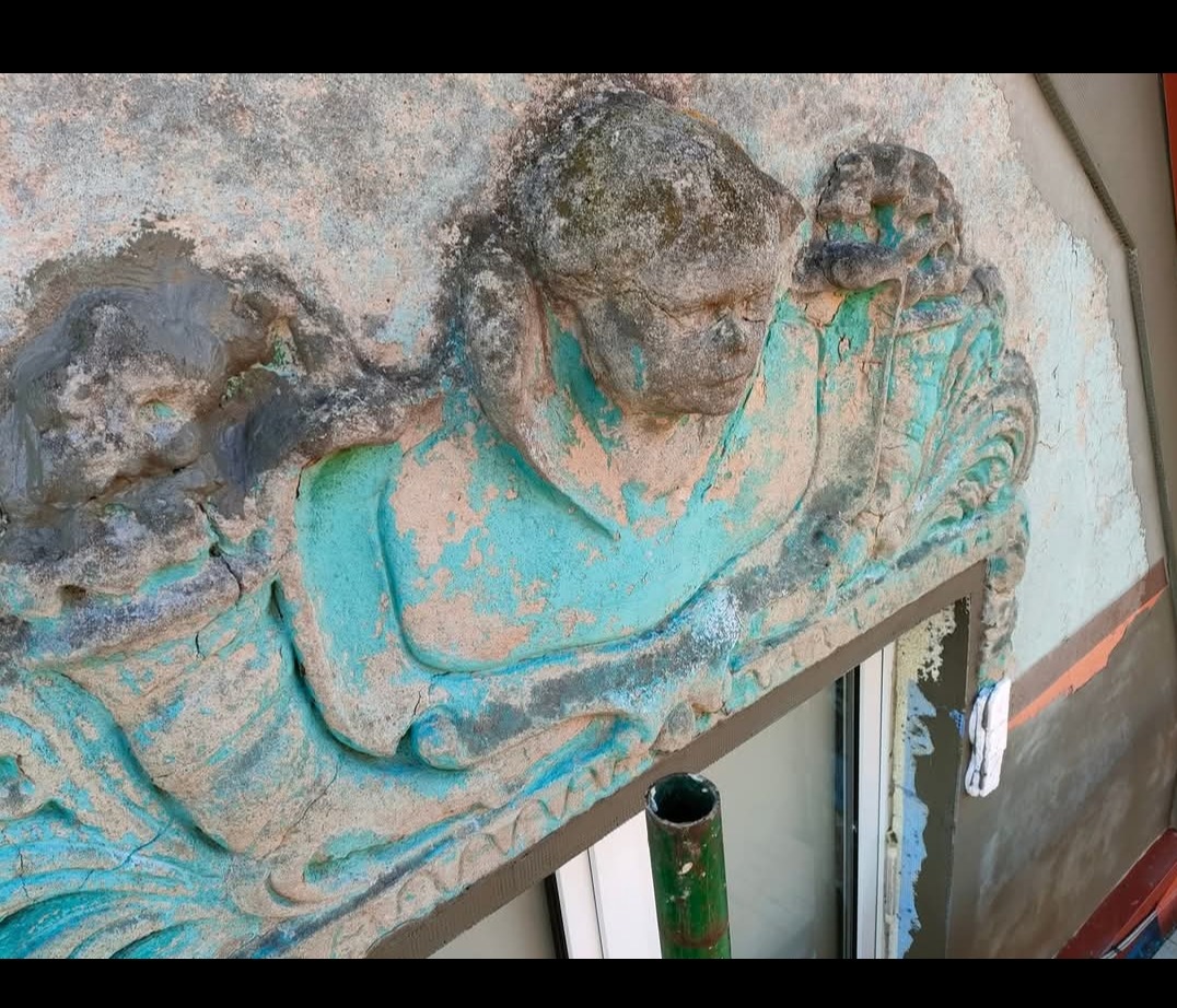 Z bliska: dekoracyjny element elewacji z rzeźbioną postacią, pokryty łuszczącą się farbą w odcieniach turkusu i szarości, nad oknem w trakcie prac renowacyjnych.