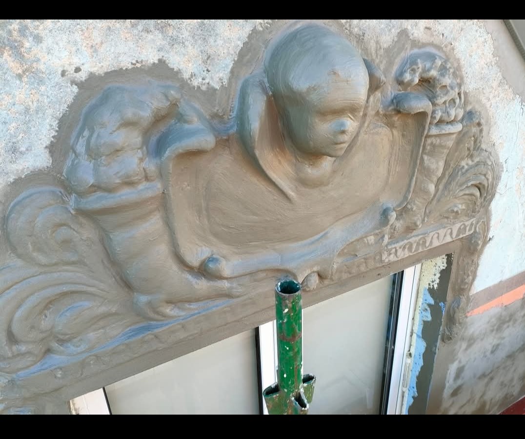 Dekoracyjne szpachlowanie elewacji nad oknem z motywem anioła i ornamentami, widoczne ślady świeżego tynku i rusztowanie. Detal wykończenia.