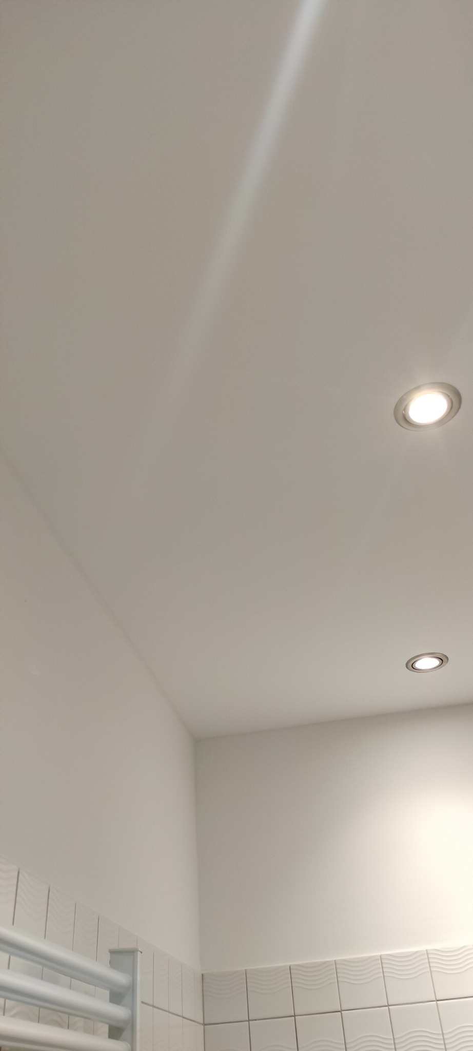 Biały sufit z dwoma wbudowanymi punktami świetlnymi, fragment ściany i grzejnika łazienkowego w dolnej części kadru. Minimalistyczne wykończenie łazienki.