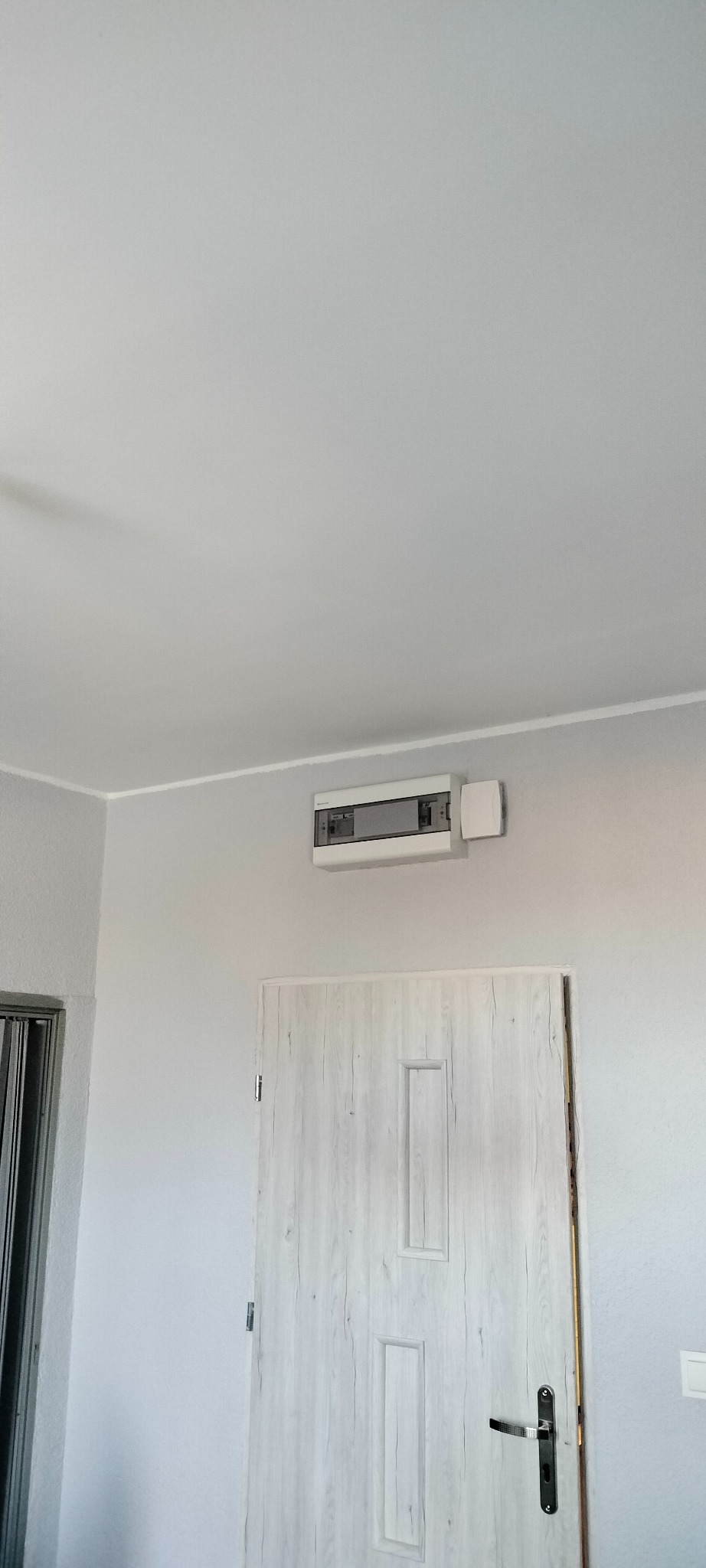 Białe drzwi z panelami i szara ściana z zamontowaną skrzynką elektryczną w jasnym wnętrzu. Minimalistyczny design, neutralna kolorystyka.