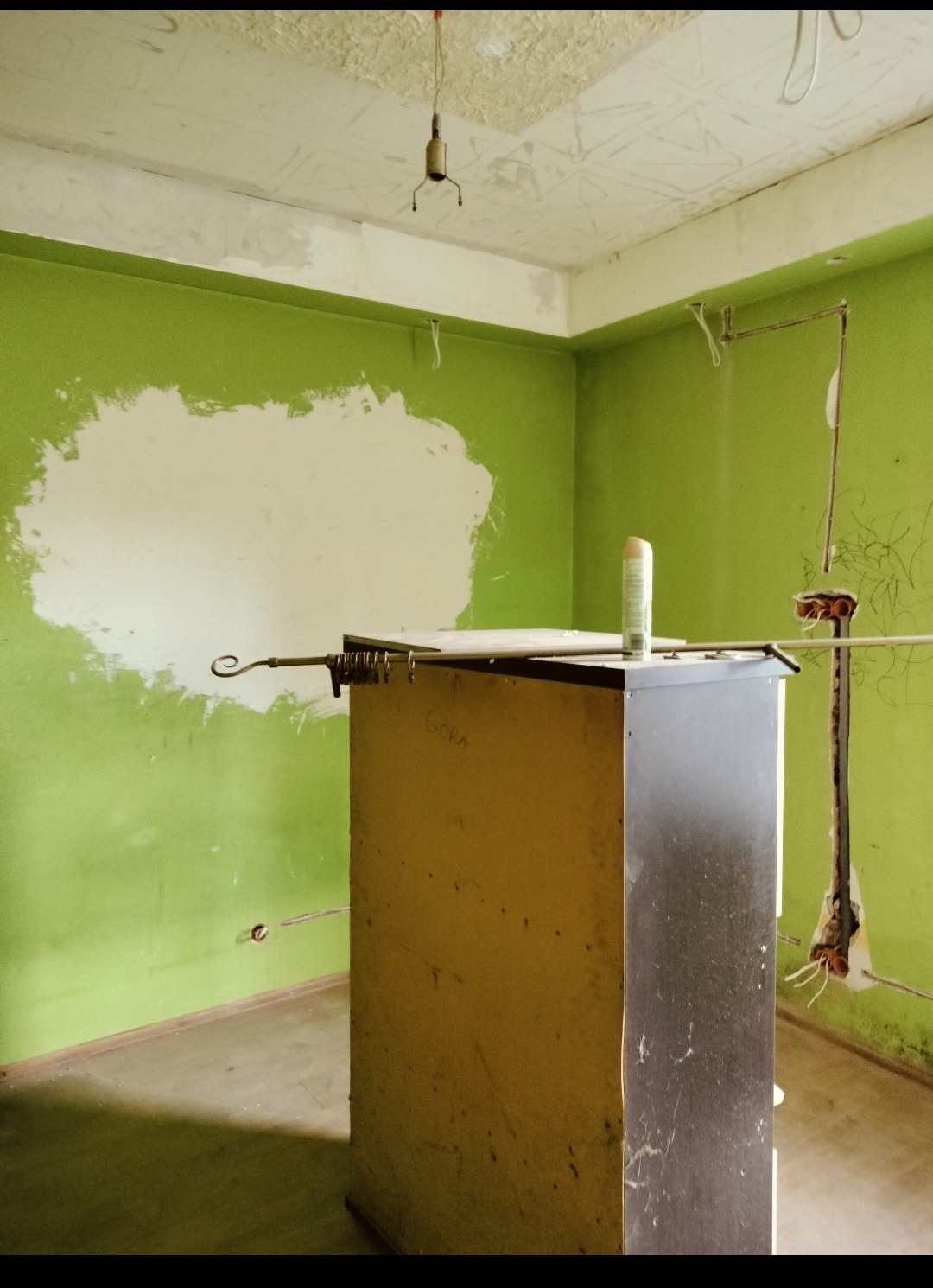 Pokój w trakcie remontu: zielone ściany z fragmentami szpachli, widoczne instalacje elektryczne, prowizoryczne oświetlenie i mebel z napisem 'GORA'.
