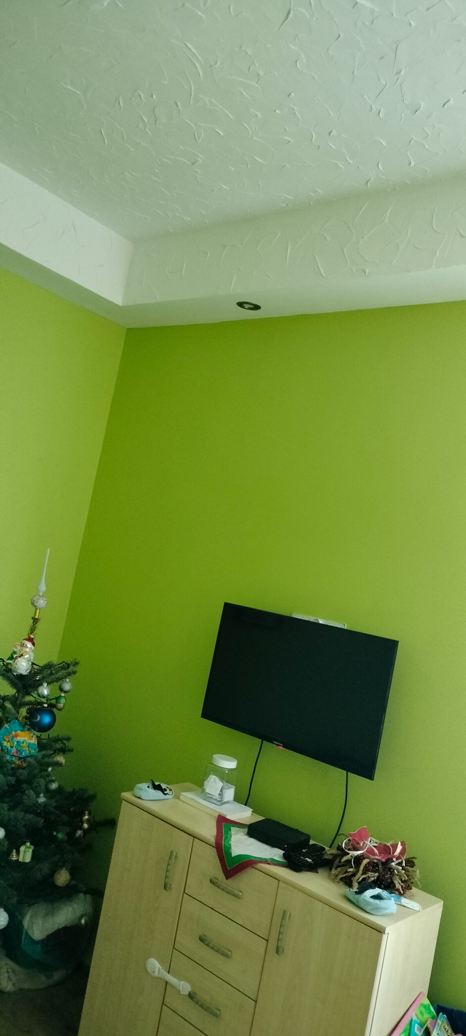 Jasnozielony pokój z telewizorem na ścianie i choinką w rogu. Sufit z fakturą, szafka pod telewizorem z dekoracjami.