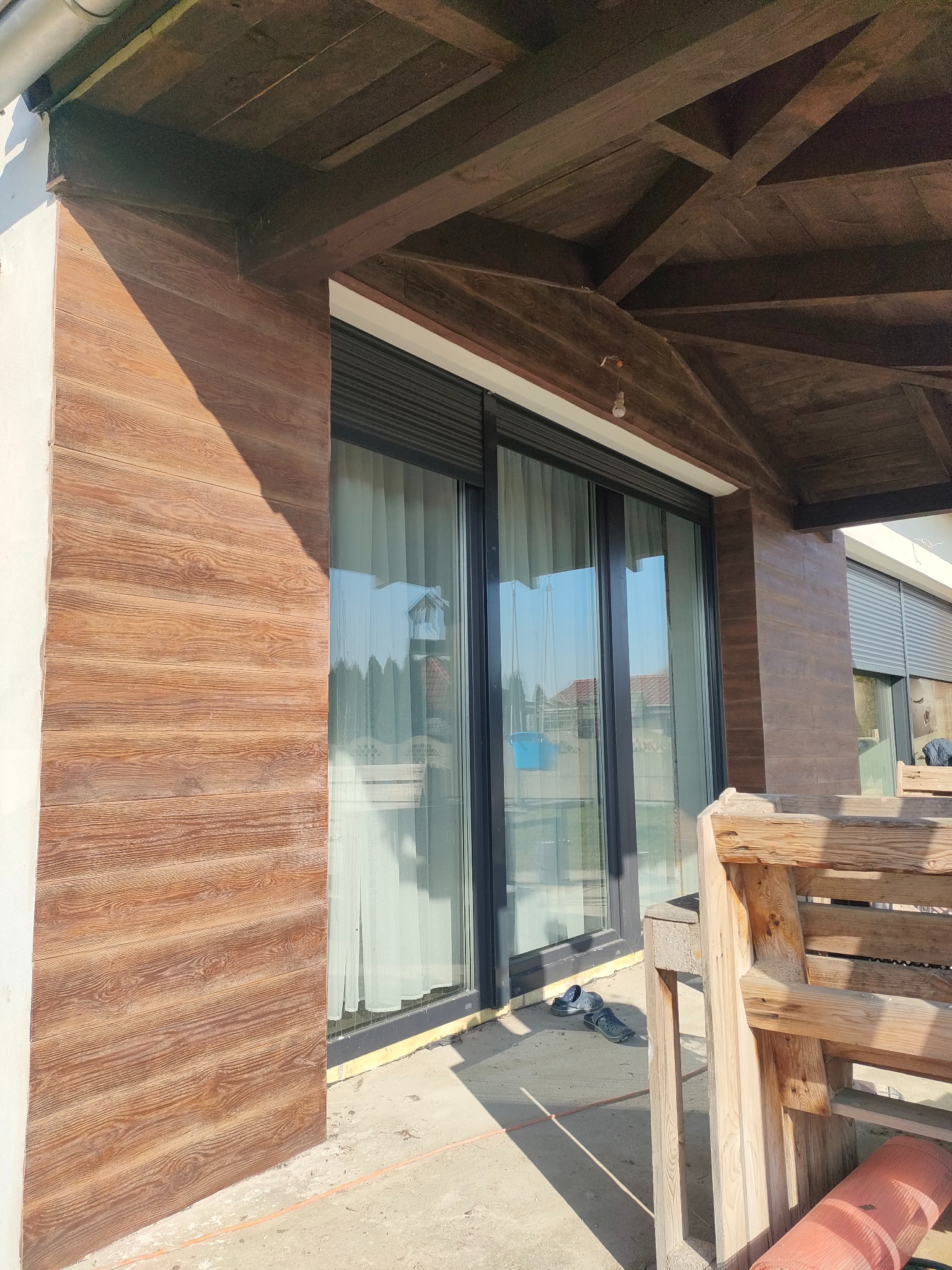 Elewacja domu z drewnopodobną okładziną, widoczne przesuwne okna z roletami zewnętrznymi i drewniana konstrukcja zadaszenia tarasu.