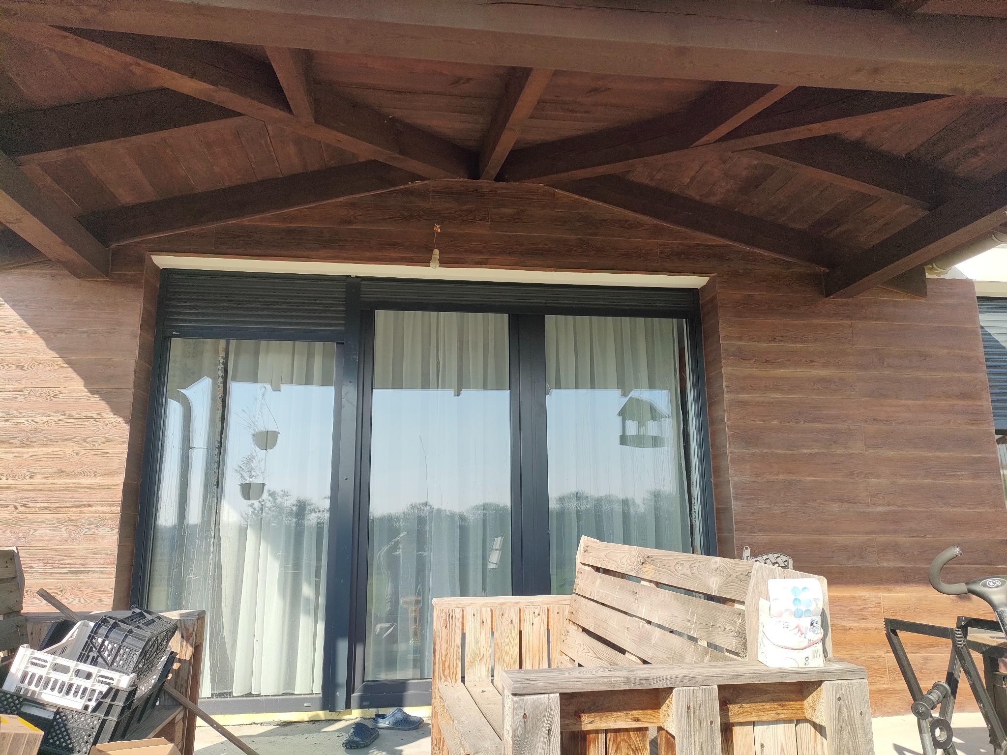 Drewniana elewacja domu z dużym oknem i zadaszeniem, widoczne elementy dekoracyjne i meble ogrodowe z palet. Ujęcie z dołu, jasne światło dzienne.