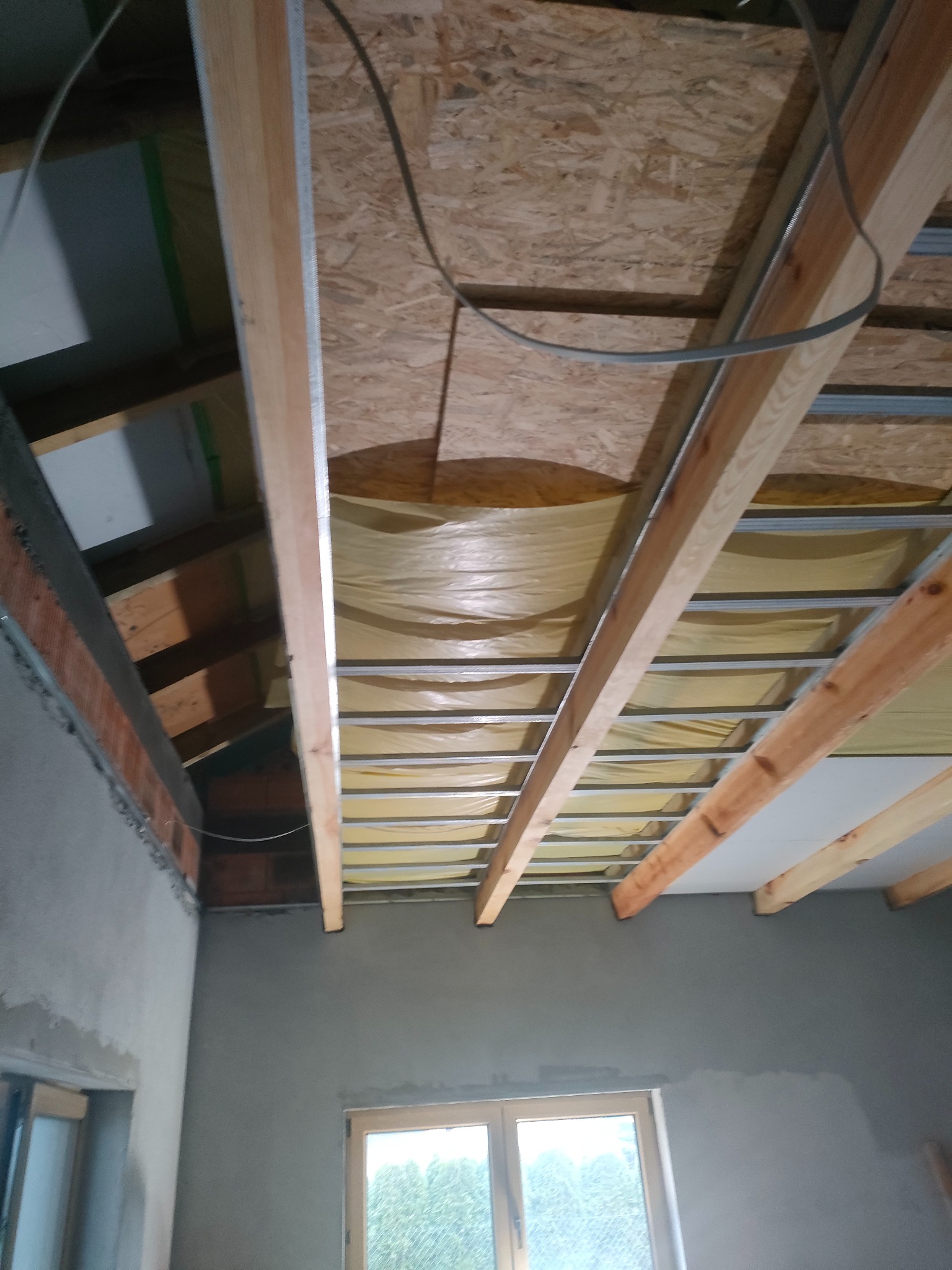 Sufit podwieszany z płyt karton-gips, widoczne drewniane belki i metalowa konstrukcja. Ściany w szarym tynku, okno w dolnej części kadru.