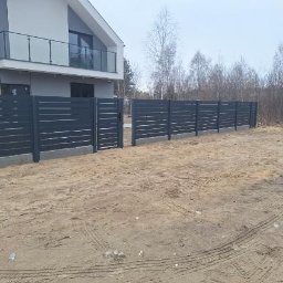 Tani-Spaw - Nowoczesne, poziome ogrodzenie panelowe w kolorze grafitowym, z furtką, na tle domu z balkonem i drzew. Widoczne ślady opon na piaszczystym podłożu.