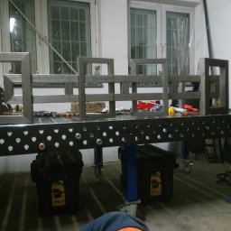 Tani-Spaw - Metalowe ramy spawane na perforowanym stole spawalniczym, widoczne narzędzia i akcesoria. W tle okna z kratami.