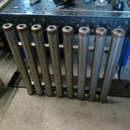 Tani-Spaw - Rząd ośmiu metalowych rur z brązowymi zakończeniami, ułożonych na szarym betonie, z blatem spawalniczym i narzędziami w tle. Widoczny hełm spawalniczy.