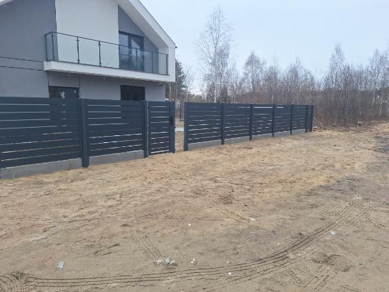 Nowoczesne, poziome ogrodzenie panelowe w kolorze grafitowym, z furtką, na tle domu z balkonem i drzew. Widoczne ślady opon na piaszczystym podłożu.