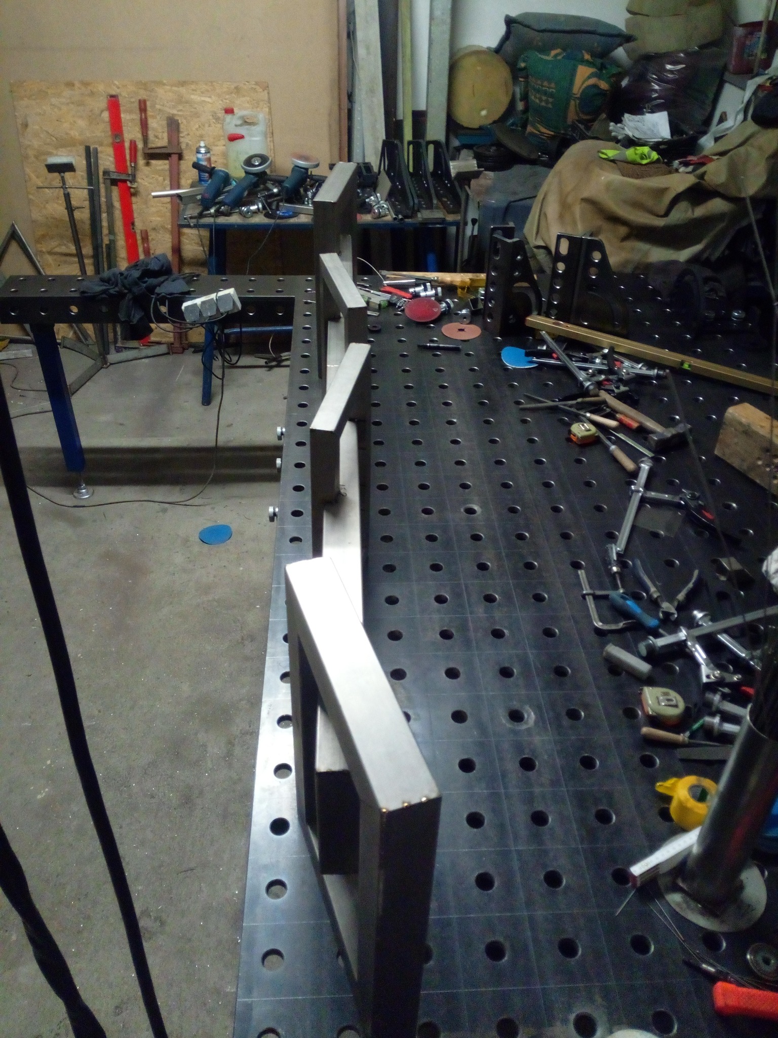 Metalowe ramy na perforowanym stole spawalniczym, narzędzia w tle. Widoczne elementy konstrukcyjne w trakcie tworzenia, warsztatowy nieład.