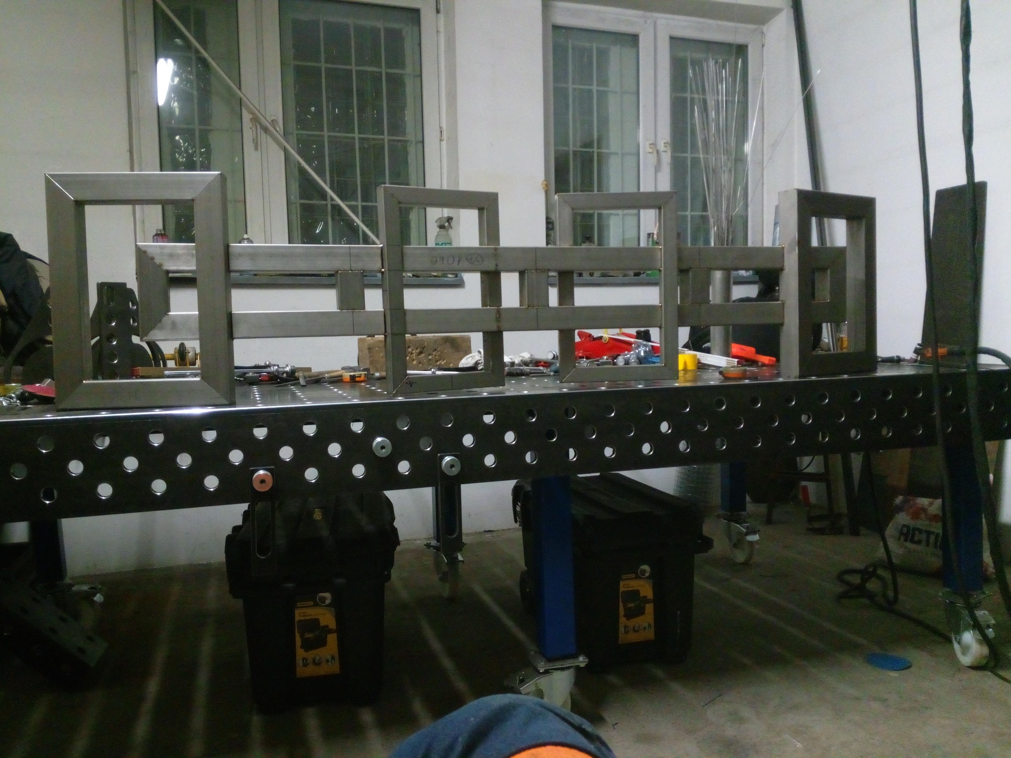 Metalowe ramy spawane na perforowanym stole spawalniczym, widoczne narzędzia i akcesoria. W tle okna z kratami.