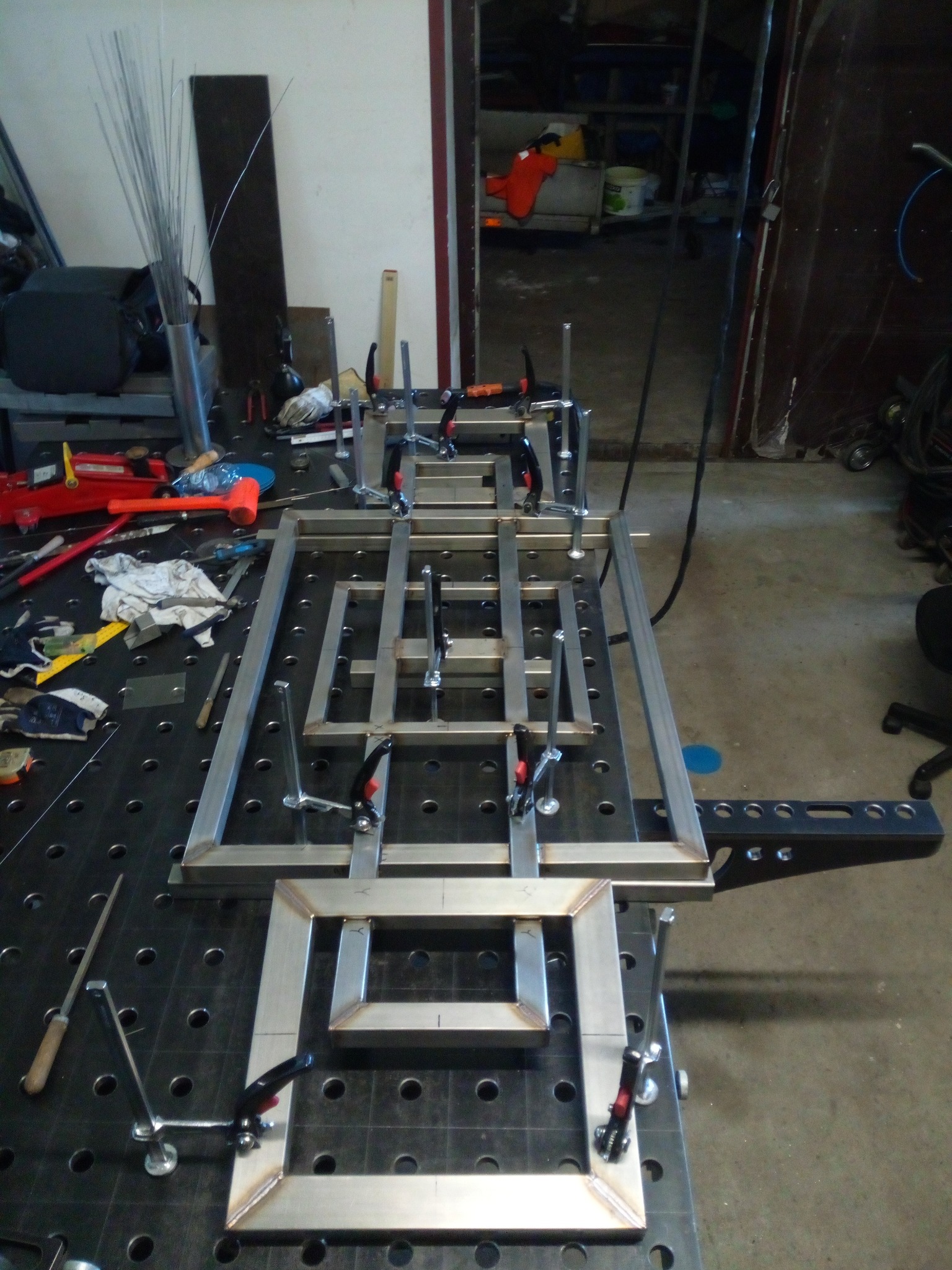 Spawanie konstrukcji metalowej z profili zamkniętych na stole spawalniczym. Elementy mocowane ściskami stolarskimi. Widoczne narzędzia i materiały spawalnicze.