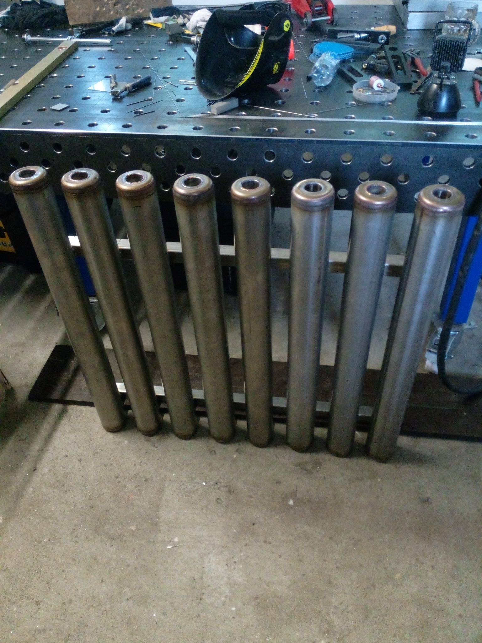 Rząd ośmiu metalowych rur z brązowymi zakończeniami, ułożonych na szarym betonie, z blatem spawalniczym i narzędziami w tle. Widoczny hełm spawalniczy.