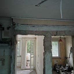 Andrii Lychko Uslugi elektryczne - Remont mieszkania w trakcie: widoczne kable elektryczne ułożone pod sufitem, ściany w stanie surowym, narzędzia i sprzęt budowlany w tle. Widok na balkon.