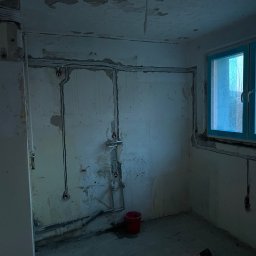 Andrii Lychko Uslugi elektryczne - Pokój w trakcie remontu z widoczną instalacją elektryczną na ścianach, czerwone wiadro na podłodze, okno z niebieską ramą. Widoczne kable i puszki elektryczne.