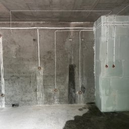 Andrii Lychko Uslugi elektryczne - Surowy betonowy pokój z widoczną instalacją elektryczną: białe kable prowadzone po ścianach i suficie, przygotowane punkty oświetleniowe. Stan surowy przed wykończeniem.