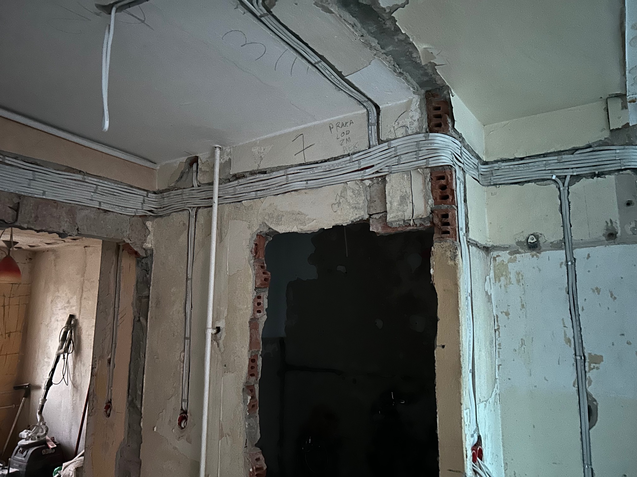 Szare kable elektryczne ułożone wzdłuż ściany i sufitu w surowym wnętrzu podczas remontu. Widoczne fragmenty cegieł i napisy na ścianie.