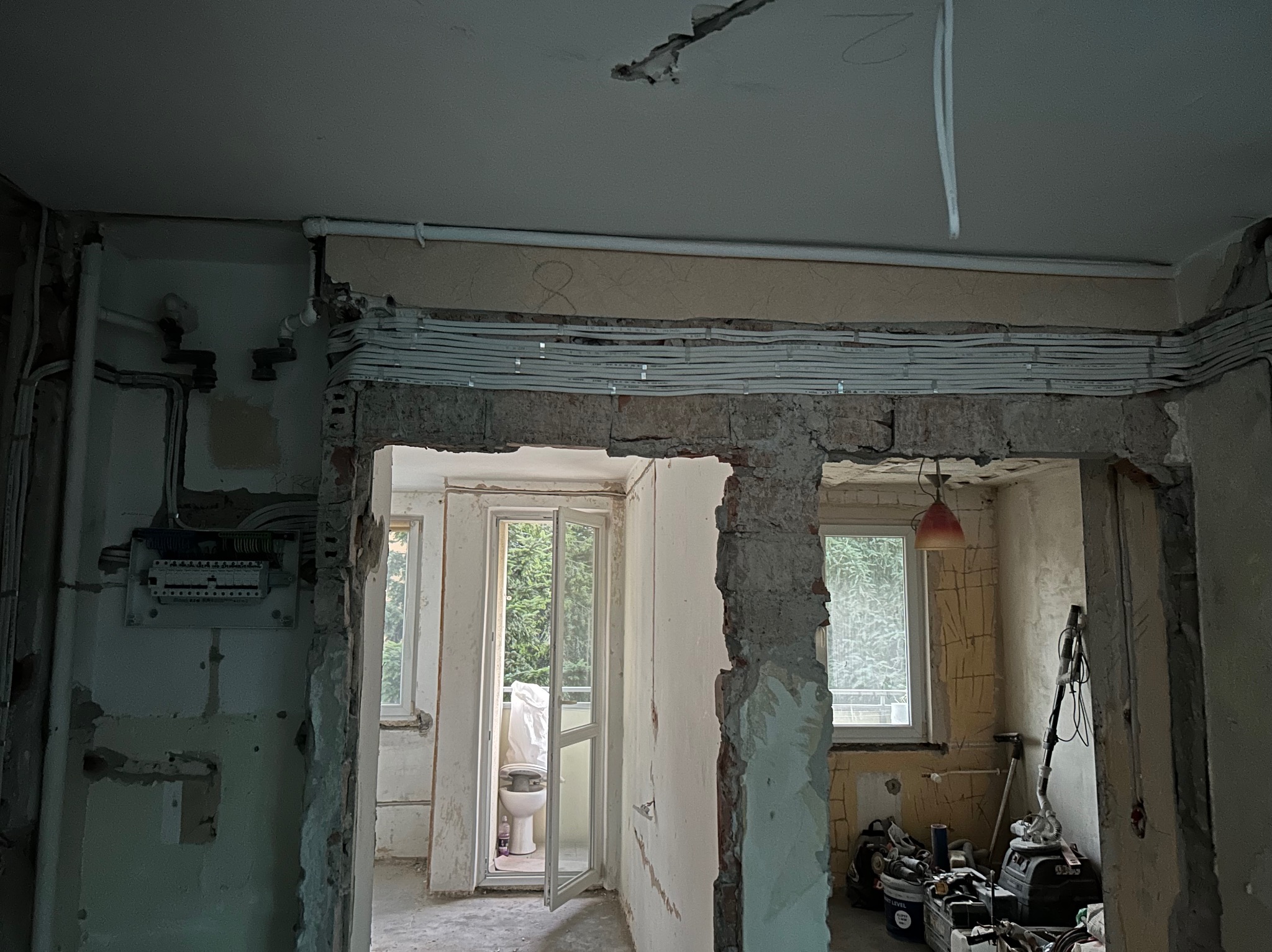 Remont mieszkania w trakcie: widoczne kable elektryczne ułożone pod sufitem, ściany w stanie surowym, narzędzia i sprzęt budowlany w tle. Widok na balkon.