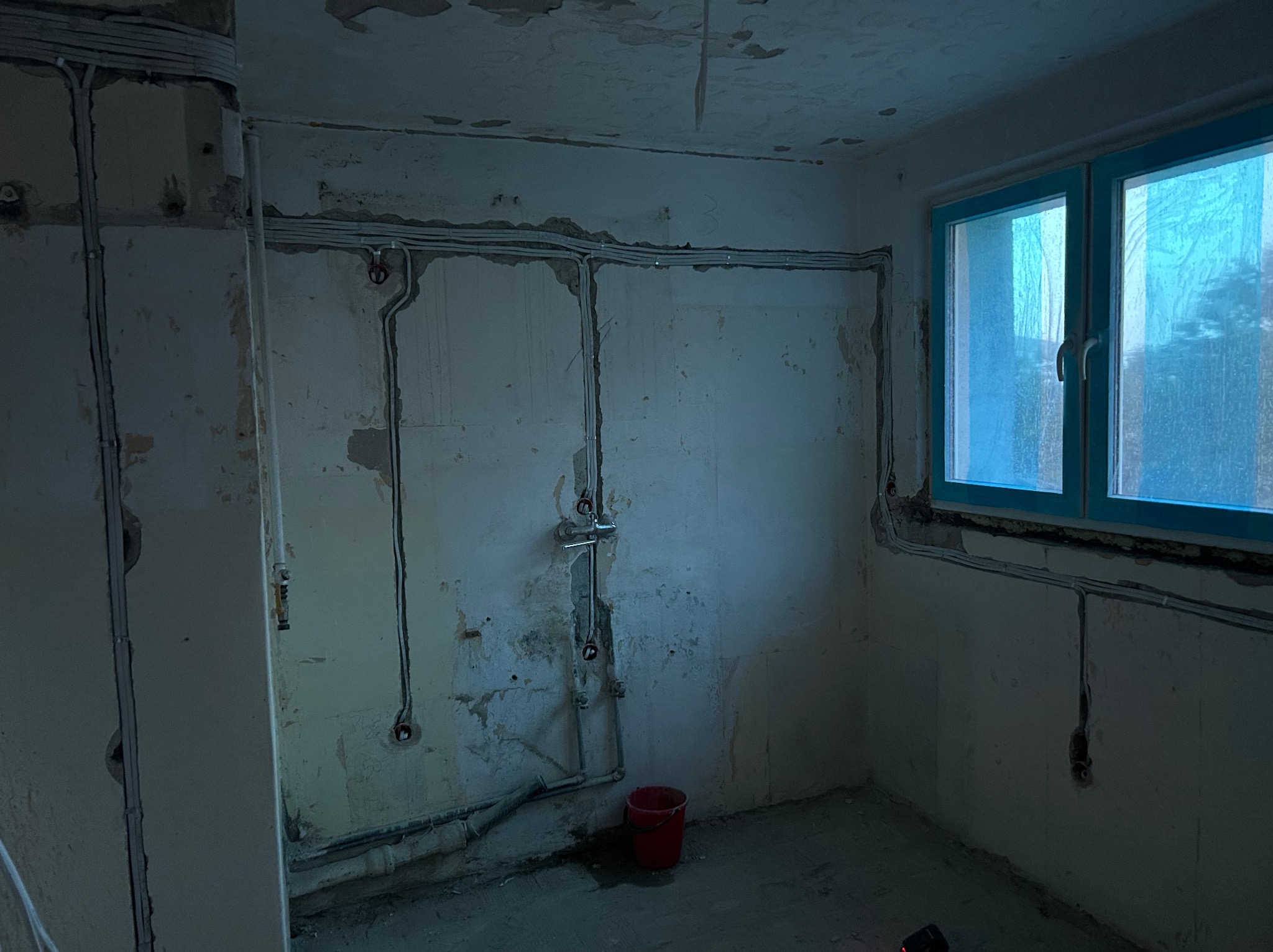 Pokój w trakcie remontu z widoczną instalacją elektryczną na ścianach, czerwone wiadro na podłodze, okno z niebieską ramą. Widoczne kable i puszki elektryczne.