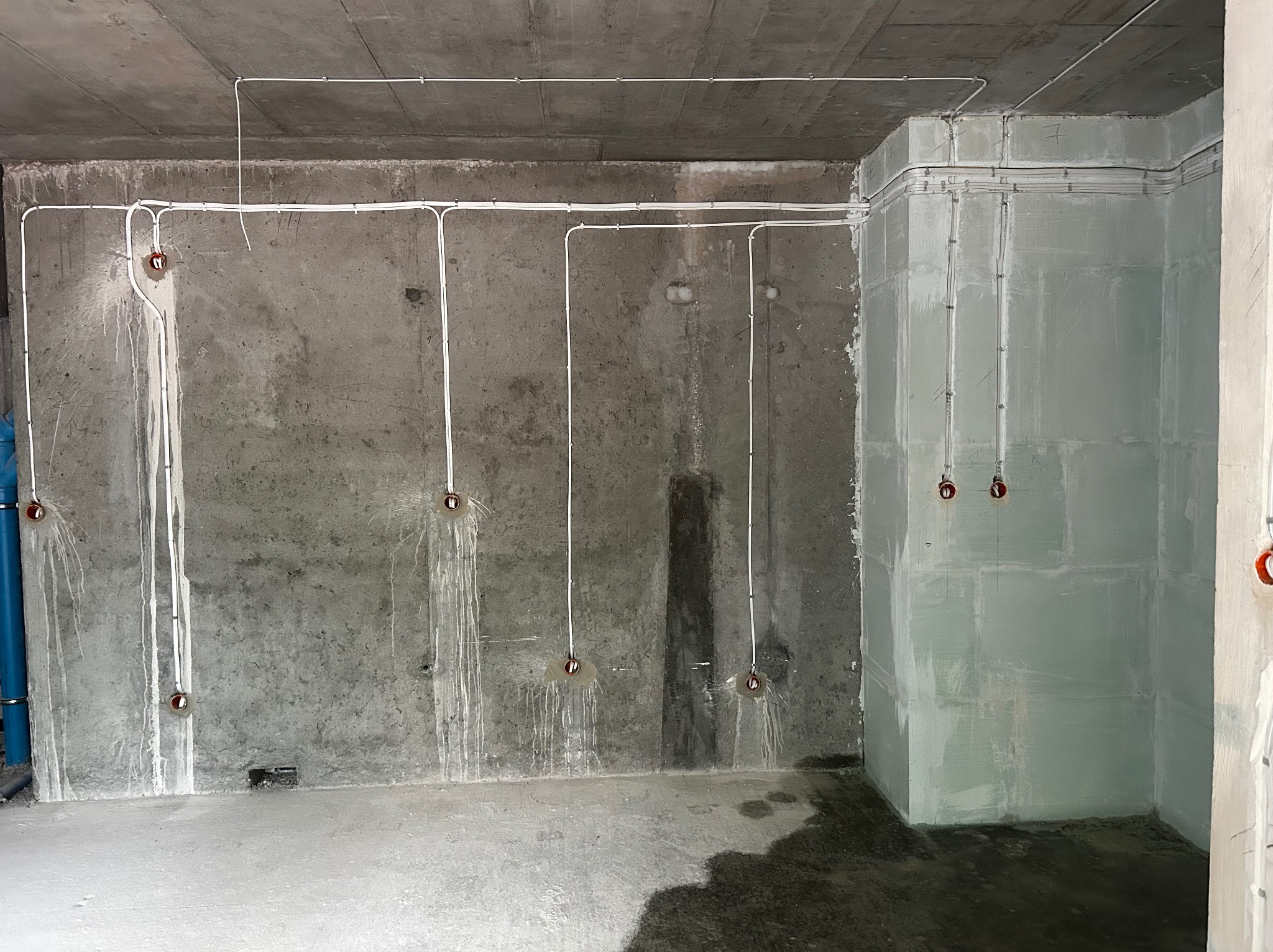 Surowy betonowy pokój z widoczną instalacją elektryczną: białe kable prowadzone po ścianach i suficie, przygotowane punkty oświetleniowe. Stan surowy przed wykończeniem.