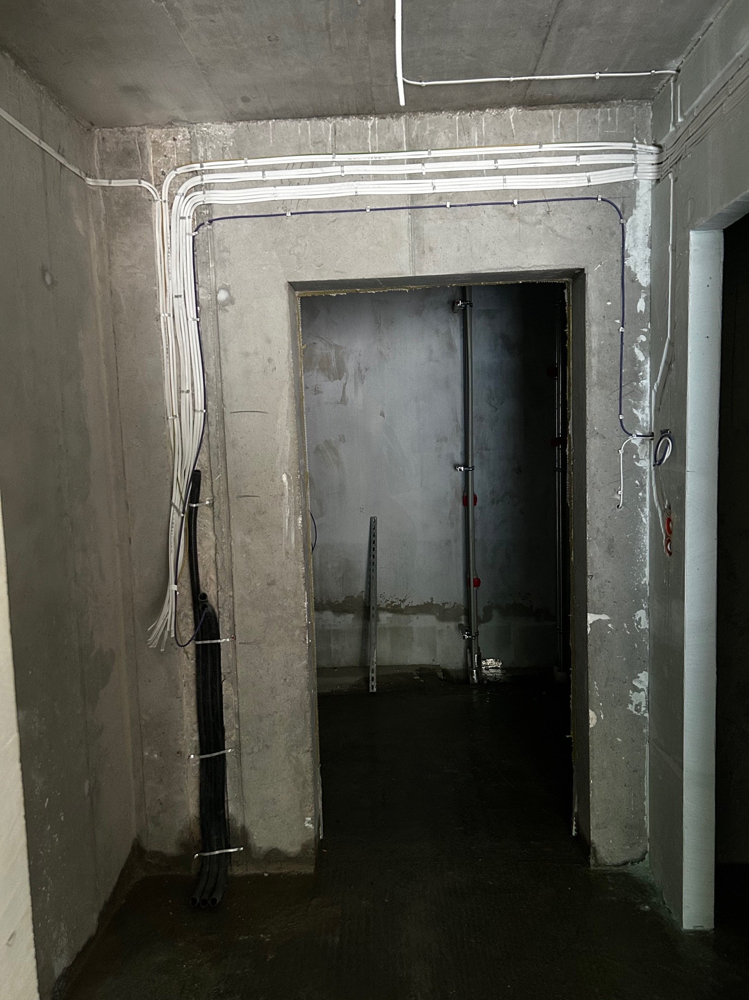 Surowy beton i instalacje elektryczne: białe kable prowadzone wzdłuż sufitu i ścian, czarne peszle pionowo, widok na surowe wnętrze przez otwór drzwiowy.