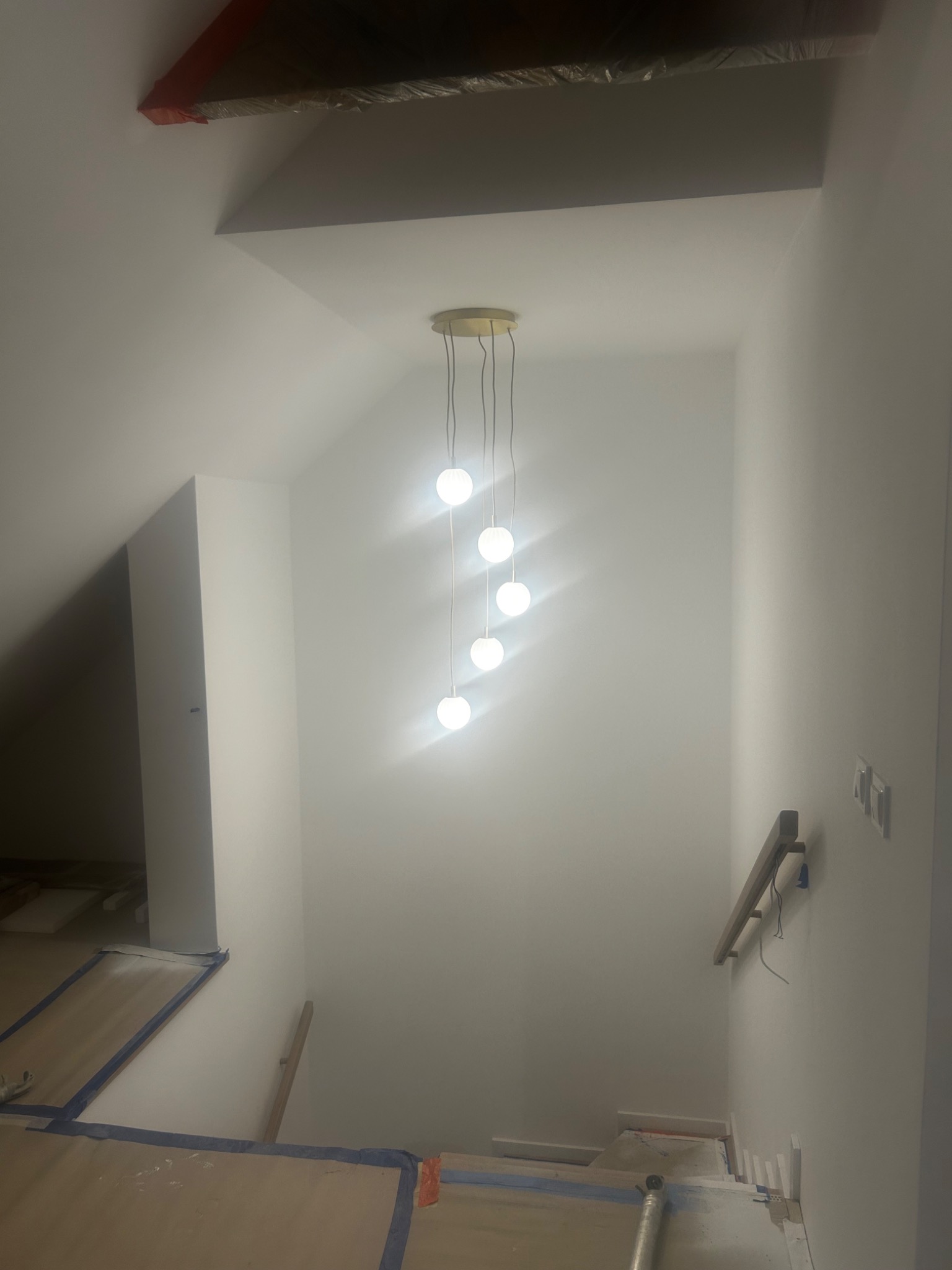 Nowoczesna lampa wisząca z kulistymi kloszami oświetla klatkę schodową w trakcie remontu. Schody zabezpieczone folią malarską, widoczne przewody elektryczne.
