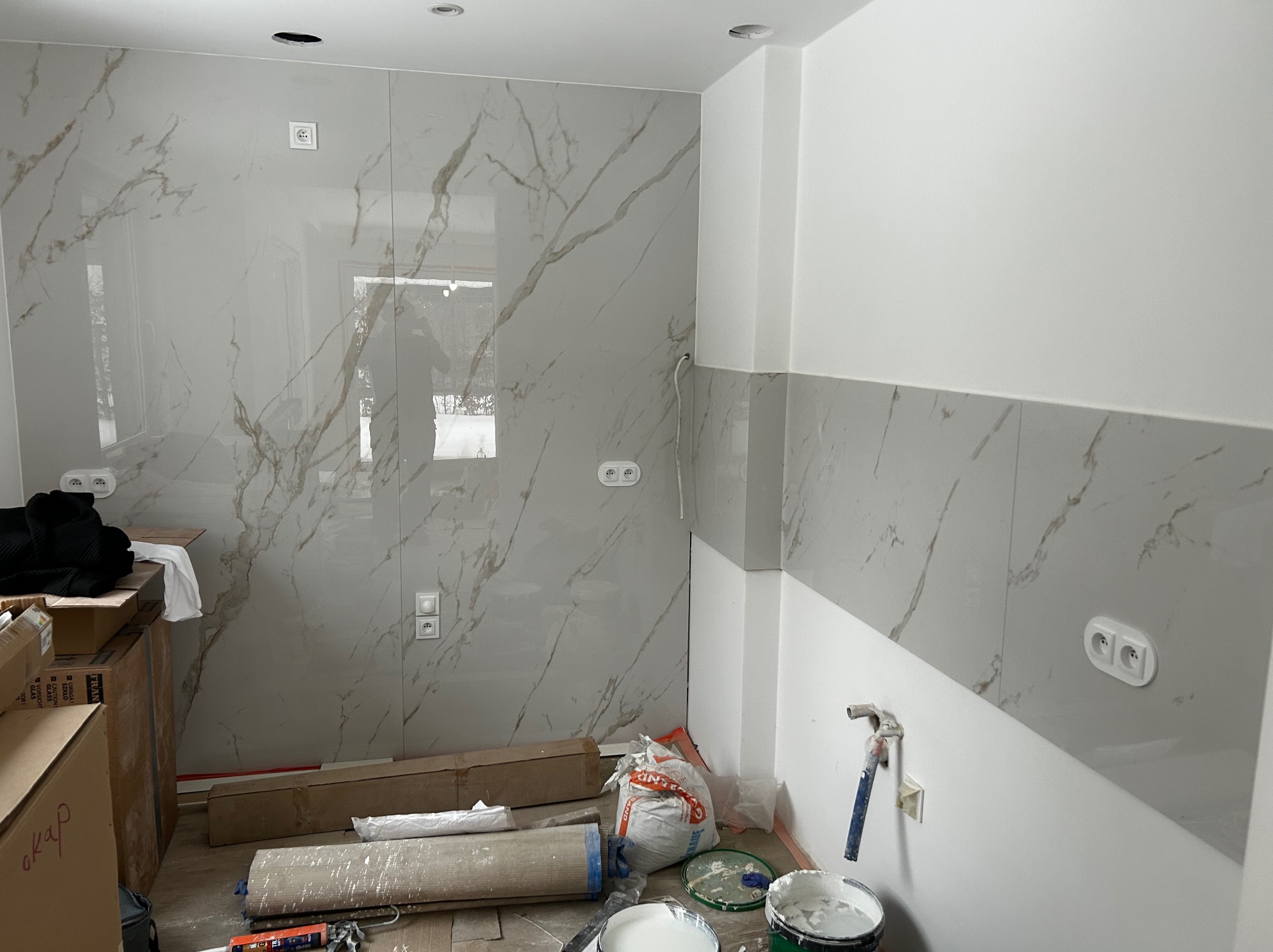 Wnętrze w trakcie wykańczania: marmurowe płytki na ścianie, gniazdka elektryczne, puszki pod oświetlenie sufitowe. Widoczne narzędzia i materiały budowlane.