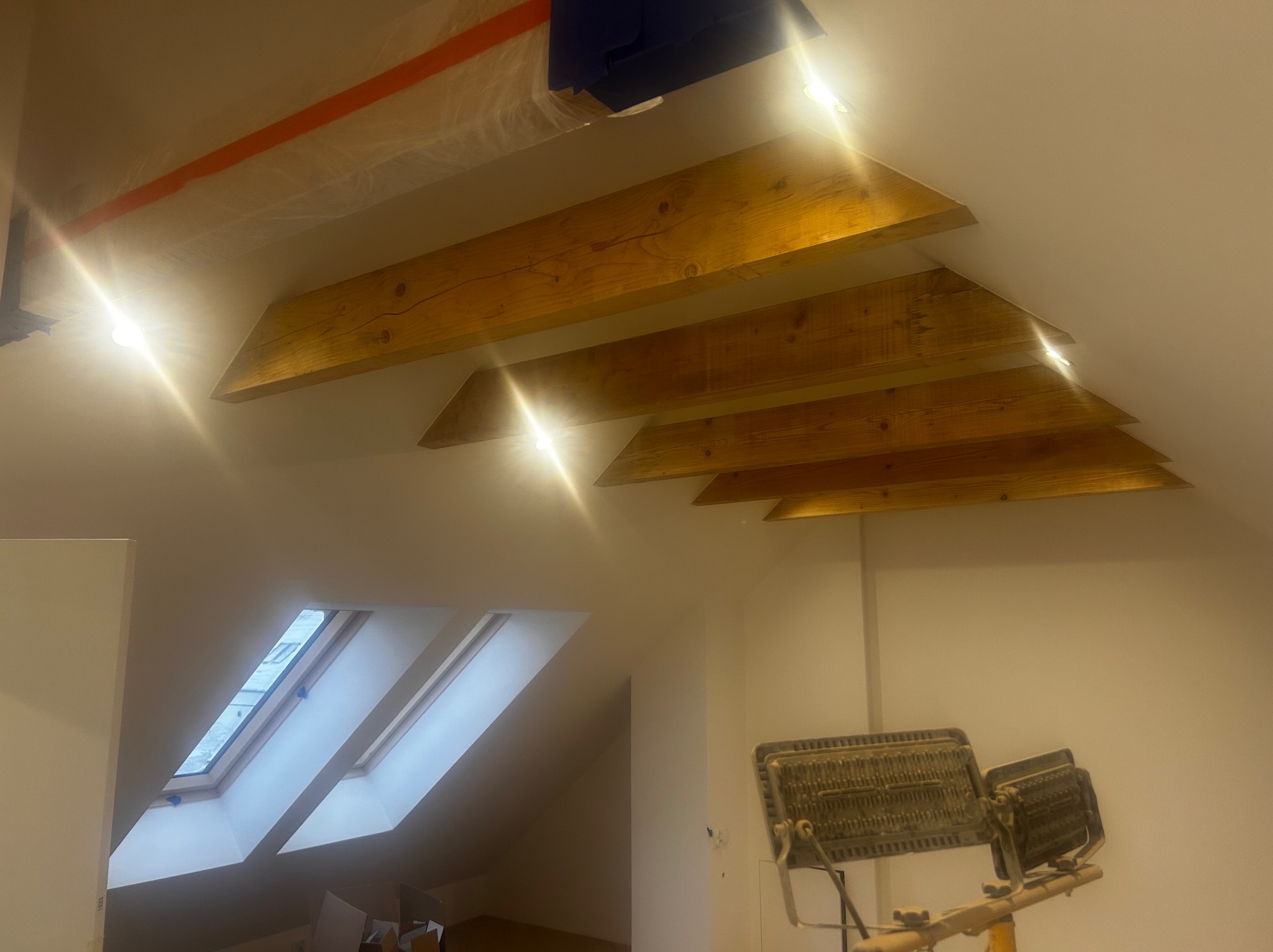 Poddasze z drewnianymi belkami i zamontowanym oświetleniem punktowym. Widoczne okno dachowe i lampa budowlana. Oświetlenie podkreśla strukturę drewna.