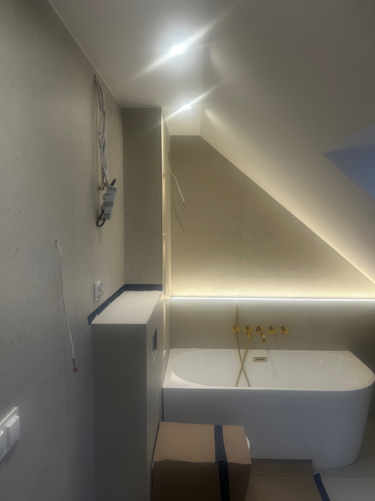 Łazienka w trakcie wykańczania: biała wanna, złota armatura, podświetlenie LED, widoczne przewody elektryczne i puszka instalacyjna na ścianie.