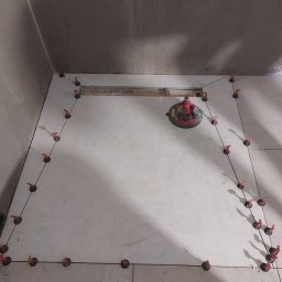 KRISTALGRESBUD VLADYSLAV BERNATSKYI - Ułożone jasne płytki podłogowe w łazience, z systemem poziomowania płytek i przyssawką do przenoszenia płytek. Widoczny odpływ liniowy.