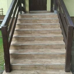 KRISTALGRESBUD VLADYSLAV BERNATSKYI - Schody wejściowe wyłożone płytkami imitującymi drewno, z ciemnobrązową balustradą, prowadzące do brązowych drzwi wejściowych. Brukowane podłoże z kratką odpływową.