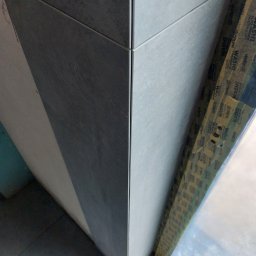 KRISTALGRESBUD VLADYSLAV BERNATSKYI - Szczegółowe ujęcie narożnika wykończonego szarymi płytkami, zabezpieczonego taśmą malarską. Widoczne precyzyjne łączenie płytek i struktura materiału.