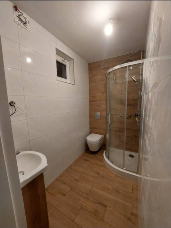 Wykończona łazienka z kabiną prysznicową, umywalką z szafką i toaletą. Białe płytki na ścianach kontrastują z drewnopodobną podłogą i ścianą za toaletą.