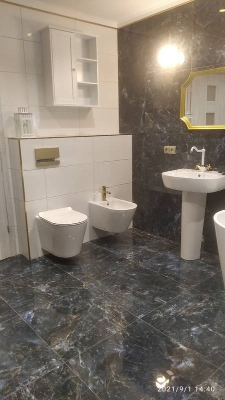 Elegancka łazienka z ciemną marmurową podłogą i białymi płytkami ściennymi. Umywalka na postumencie, toaleta i bidet w nowoczesnym stylu. Lustro w złotej ramie i biała szafka.