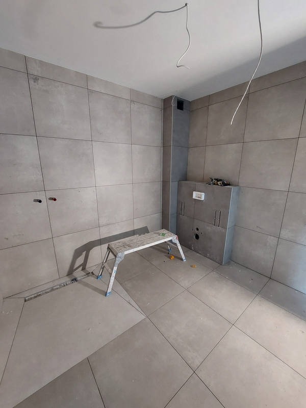 Wnętrze łazienki w trakcie wykańczania: szare płytki na ścianach i podłodze, drabinka aluminiowa, widoczne przewody elektryczne. Minimalistyczny styl, surowy klimat.