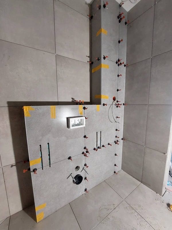 Szare płytki w łazience w trakcie montażu, z użyciem systemu poziomowania płytek i taśmy maskującej. Widoczne otwory na instalacje i biała skrzynka podtynkowa.