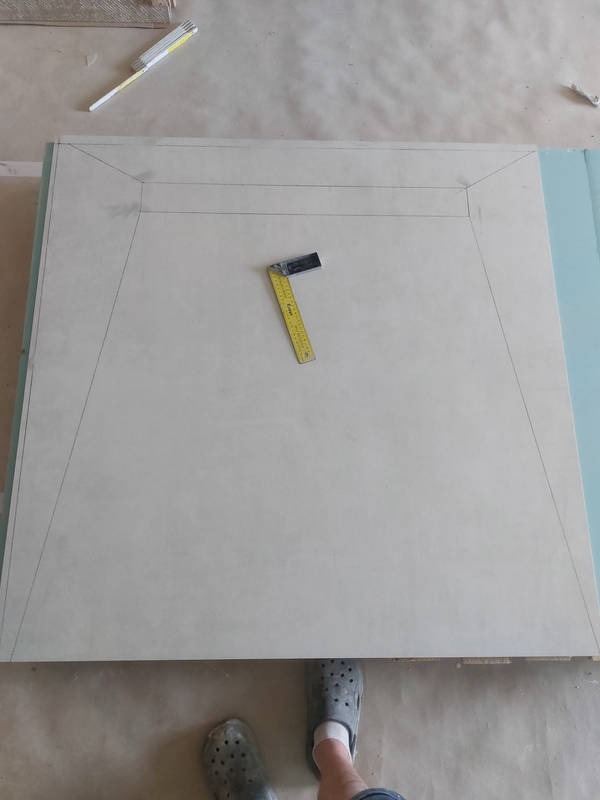 Płyta gipsowa z odrysowanym trapezoidalnym kształtem, kątownik stolarski i miarka na wierzchu, przygotowana do cięcia, widok z góry.