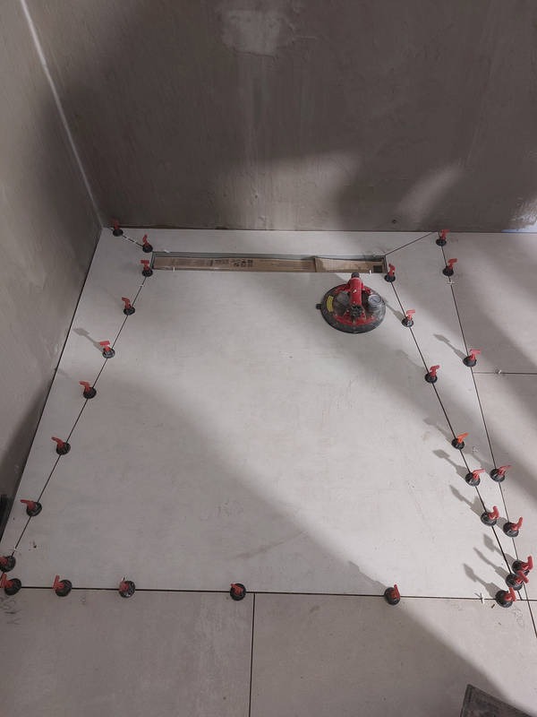 Ułożone jasne płytki podłogowe w łazience, z systemem poziomowania płytek i przyssawką do przenoszenia płytek. Widoczny odpływ liniowy.