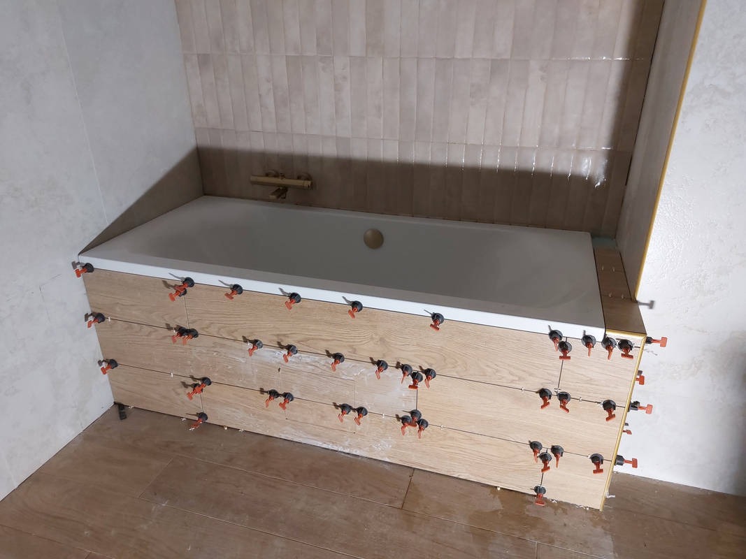 Biała wanna obłożona płytkami imitującymi drewno, z systemem poziomowania płytek. Na ścianie i podłodze również płytki w odcieniach beżu i brązu. Widok na fragment wykończonej łazienki.