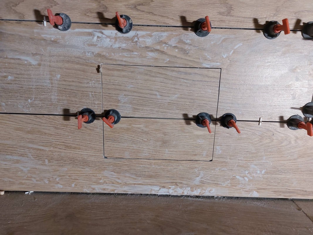 Układanie płytek imitujących drewno z użyciem systemu poziomowania. Widoczny otwór rewizyjny i resztki kleju. Detal z bliska.