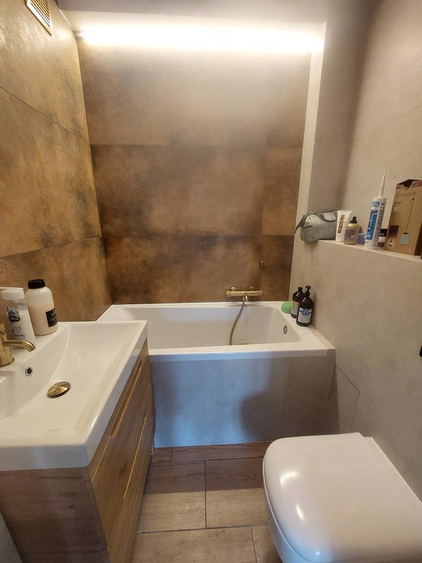 Nowoczesna łazienka z wanną, umywalką z szafką z drewnianymi frontami i toaletą. Brązowe płytki na ścianie w strefie wanny, oświetlenie LED.