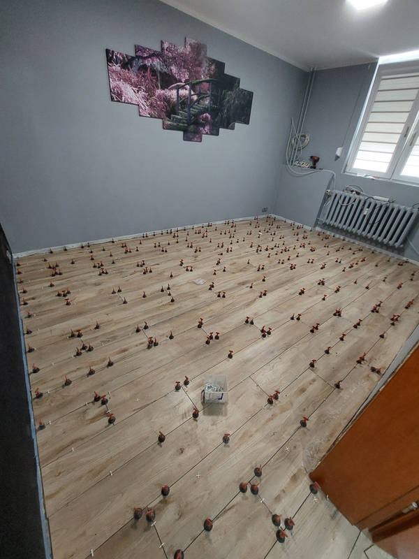 Układanie paneli podłogowych w pokoju z szarą ścianą i obrazem. Widoczne klipsy poziomujące i skrzynka z narzędziami. Jasne panele ułożone w jodełkę.