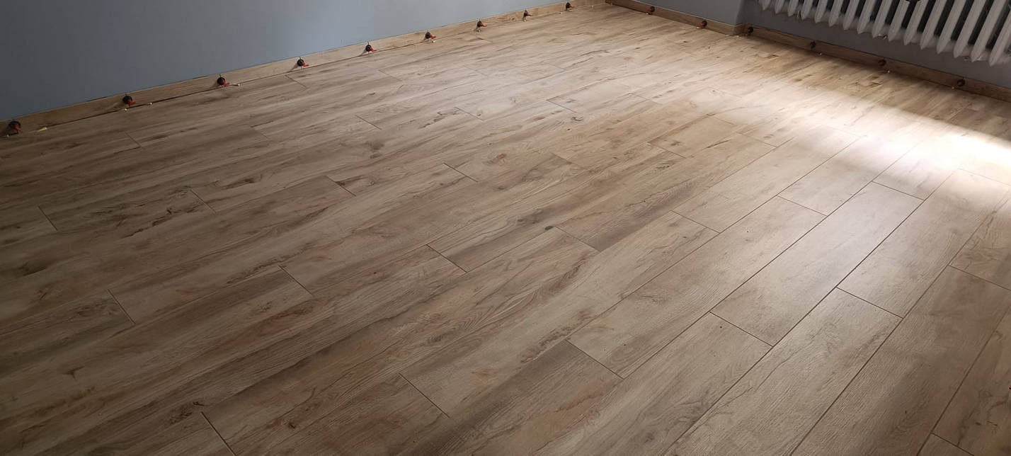 Ułożone pod kątem płytki imitujące drewno, z widocznymi elementami poziomowania przy ścianach i grzejniku, w jasnym, naturalnym oświetleniu.