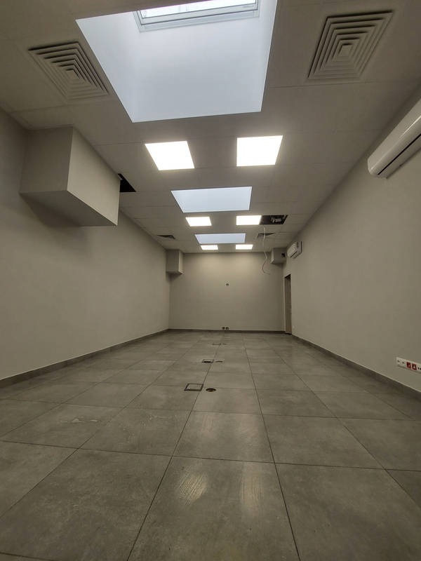 Wykończone wnętrze biurowe z szarą podłogą z płytek, białymi ścianami i sufitem z wbudowanym oświetleniem. Widoczne kratki wentylacyjne i klimatyzacja.