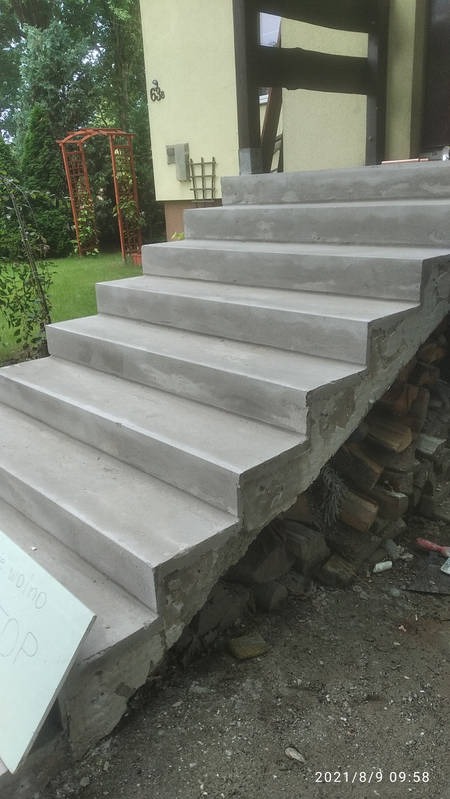 Betonowe schody zewnętrzne prowadzące do domu, widoczne z boku, z elementami ogrodu w tle. Widoczne ślady prac budowlanych.