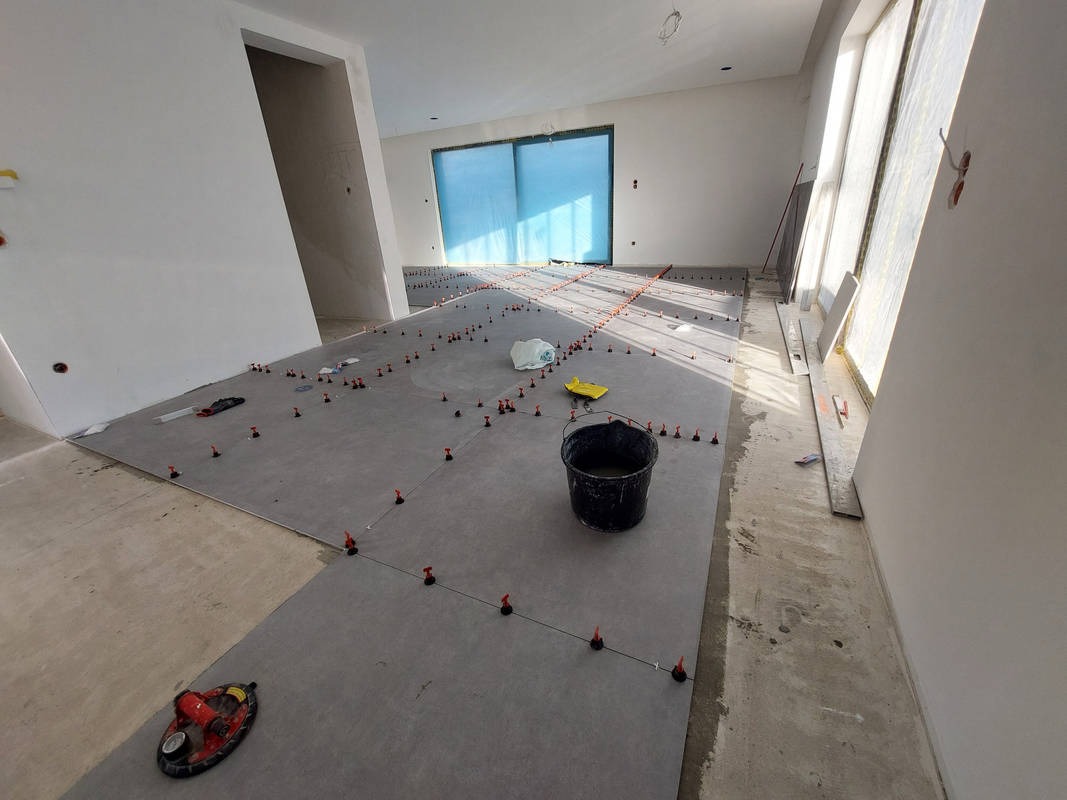 Szare płytki podłogowe w trakcie układania, z systemem poziomowania, wiadro i przyssawka w przestronnym wnętrzu z dużymi oknami. Widok na etap prac glazurniczych.