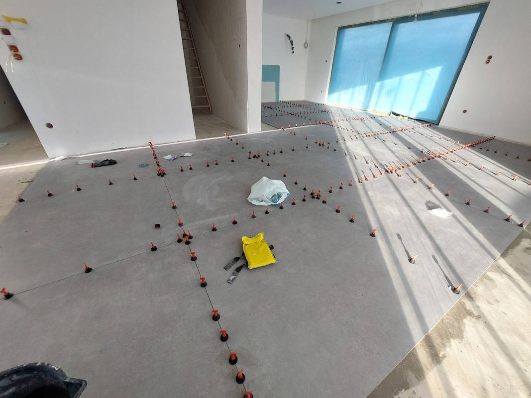 Szara podłoga z ułożonymi płytkami, system poziomowania płytek, worek i żółta torba narzędziowa. W tle drabina i okno. Układanie płytek w przestronnym wnętrzu.