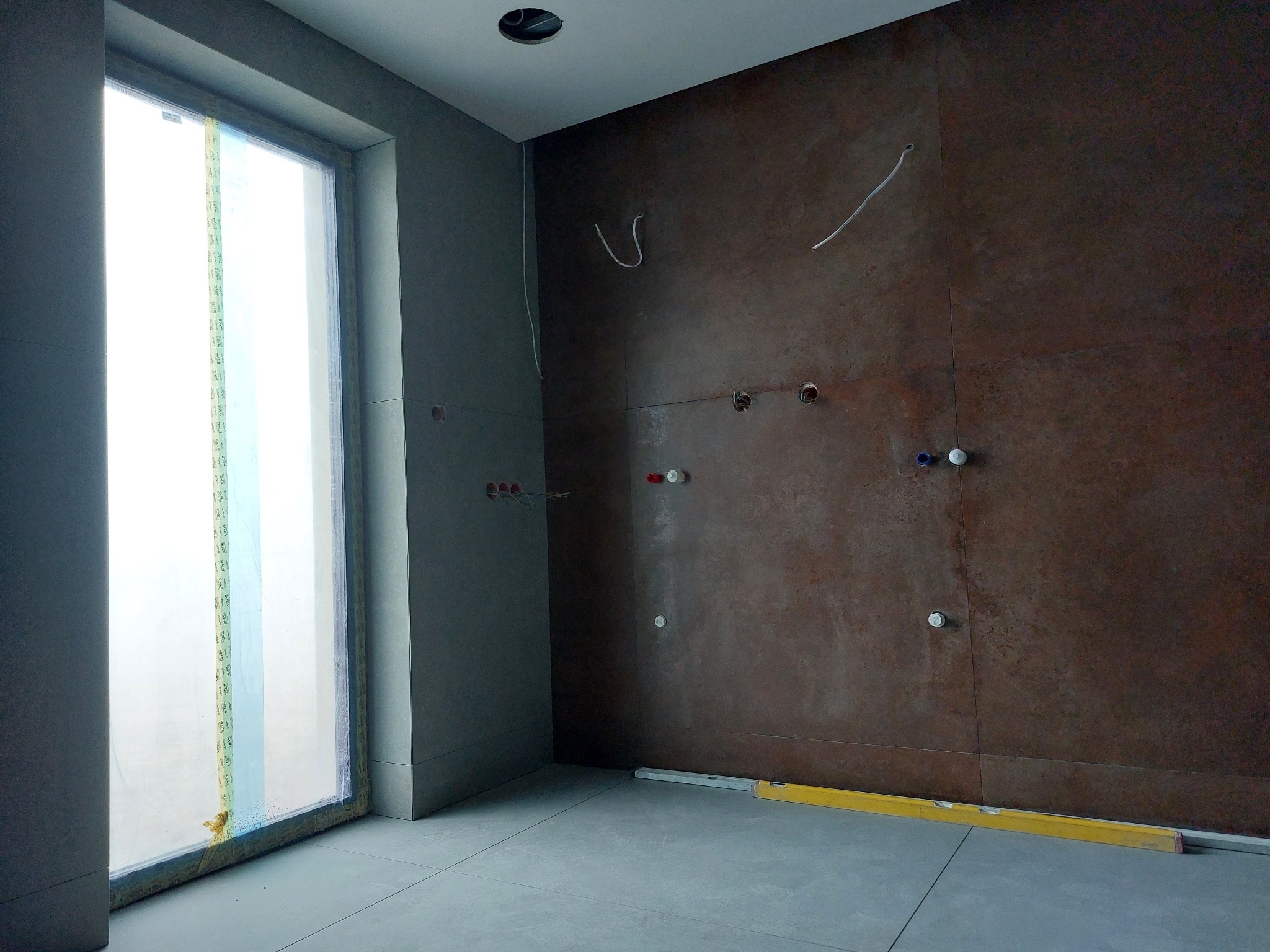 Wnętrze łazienki w trakcie wykańczania: szare płytki podłogowe i ścienne, brązowa ściana z instalacją wodną, okno zabezpieczone folią, poziomica na podłodze.