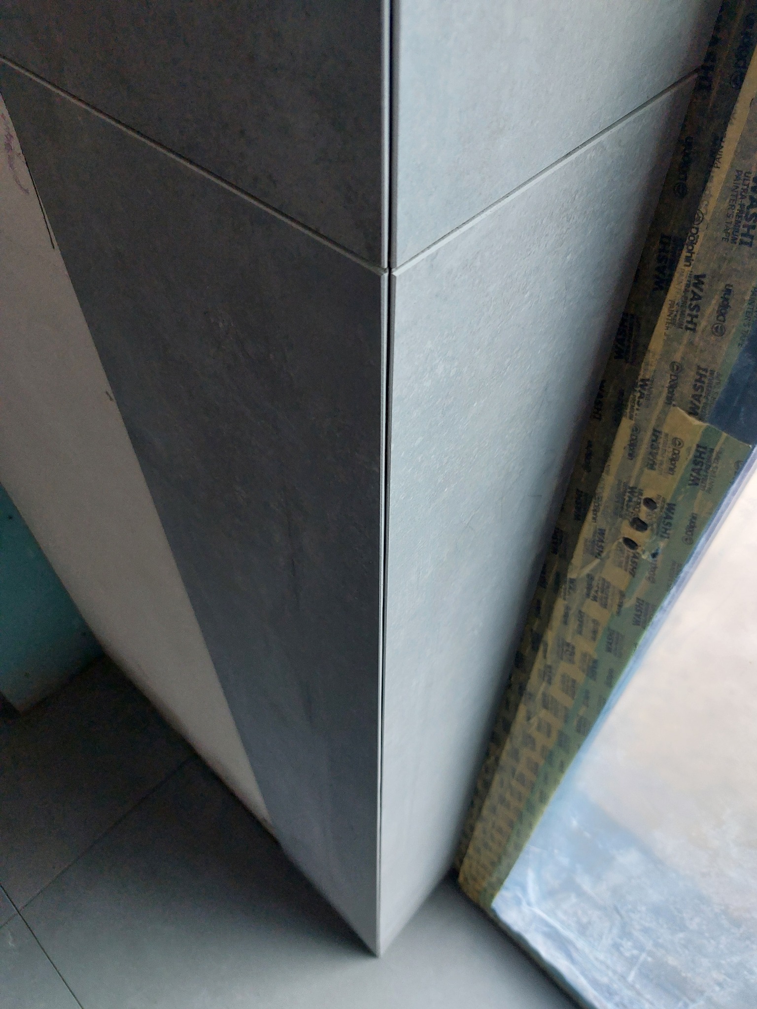 Szczegółowe ujęcie narożnika wykończonego szarymi płytkami, zabezpieczonego taśmą malarską. Widoczne precyzyjne łączenie płytek i struktura materiału.