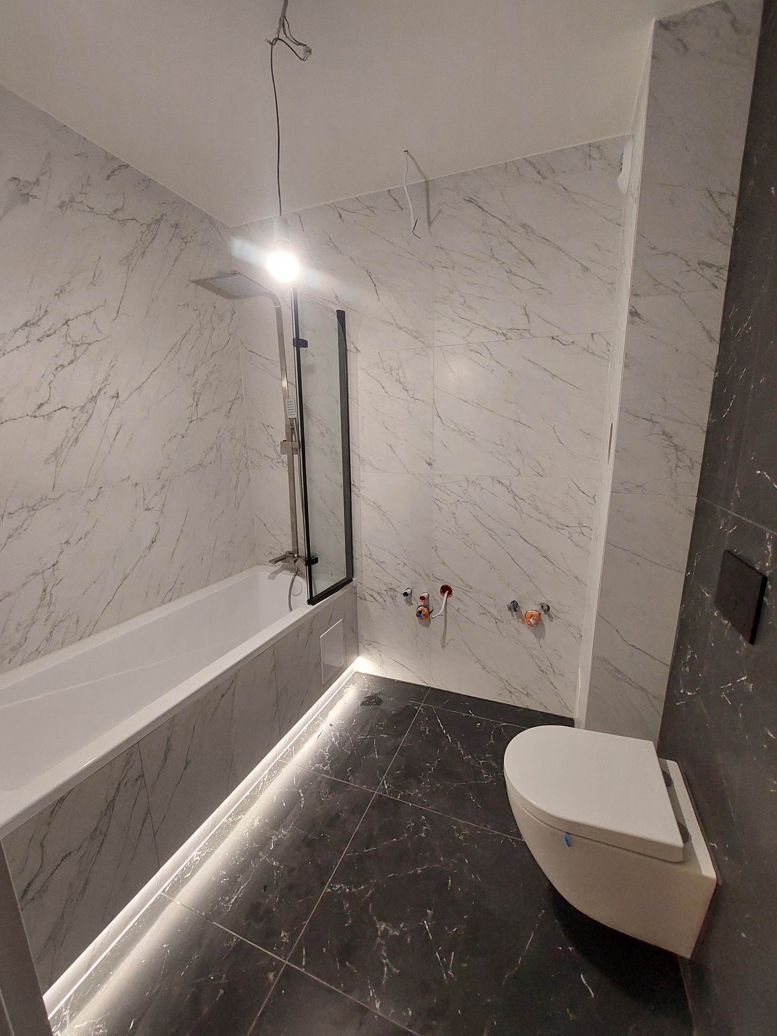 Wykończona łazienka z marmurowymi płytkami, podświetlaną wanną i podwieszaną toaletą. Widoczne przyłącza wodne. Minimalistyczny design.